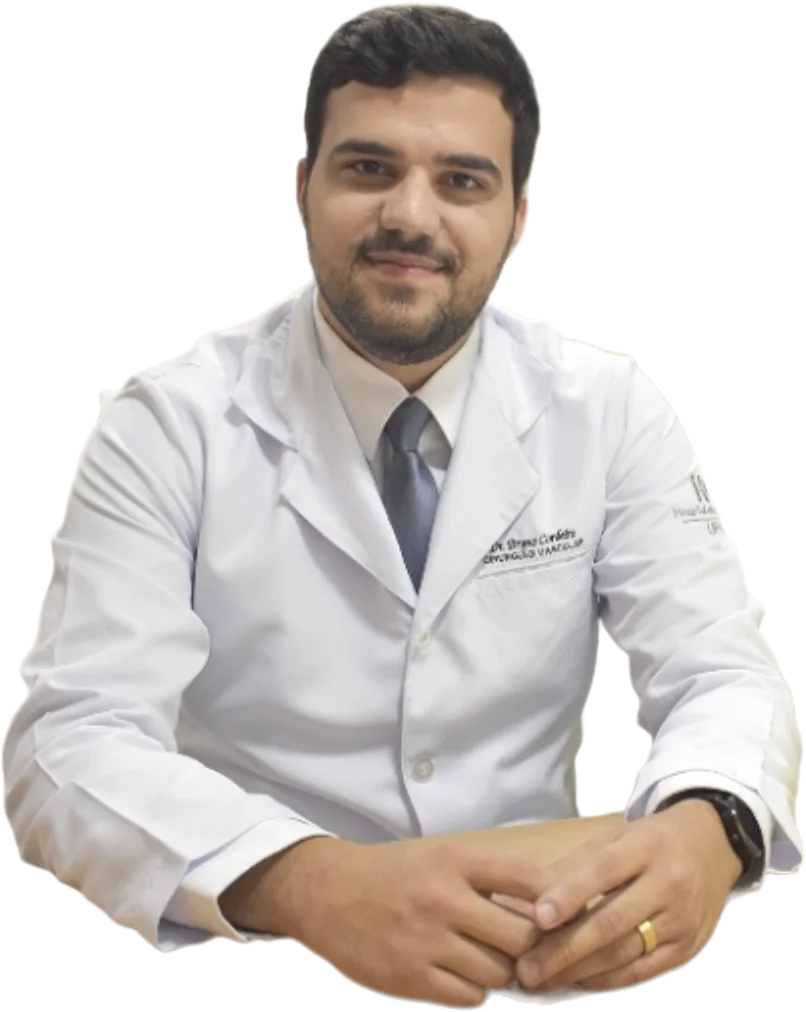 Dr. Bruno Cordeiro de jaleco branco e gravata azul, sentado à mesa do consultório, mãos juntas, olhando para a câmera.