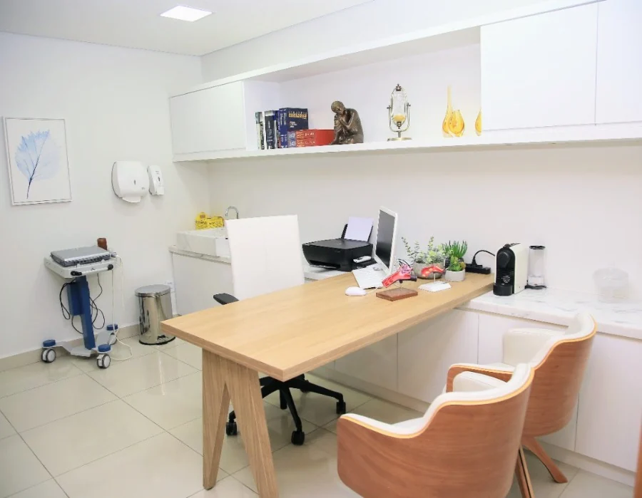 Consultório claro, com mesa de madeira, duas poltronas para o paciente e prateleiras brancas com livros.