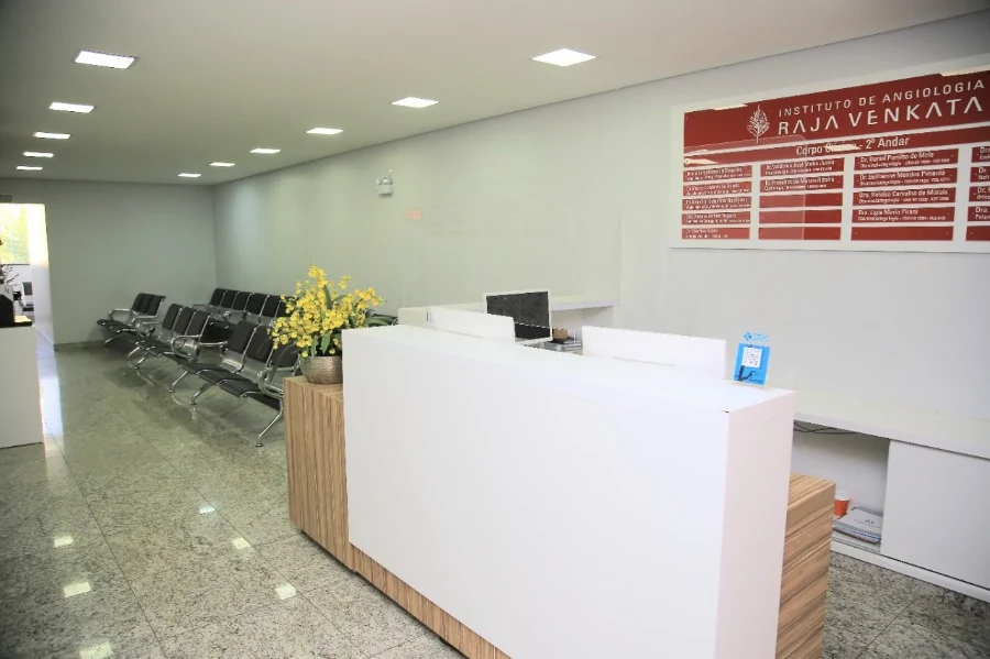 Sala de espera clara, com cadeiras em fileira, piso de granito e balcão branco com flores amarelas.